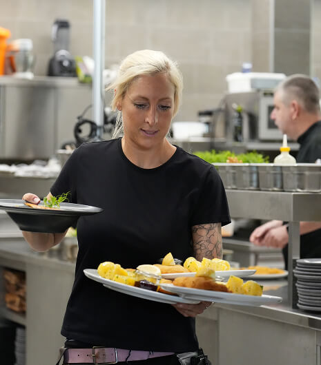 Blonde Frau trägt mehrere Teller mit Essen in Küche.