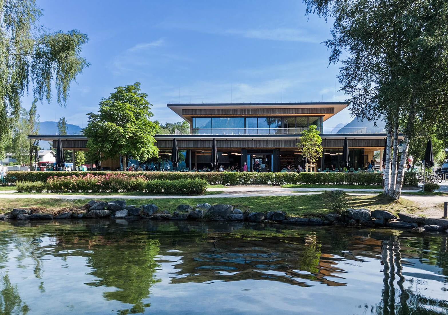 Modernes Gebäude am See mit Terrasse, Bäumen, Bergen.