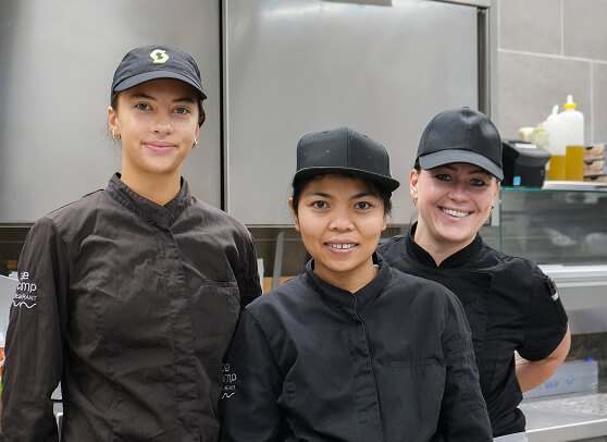 Drei lächelnde Frauen in schwarzen Kochjacken und Mützen.